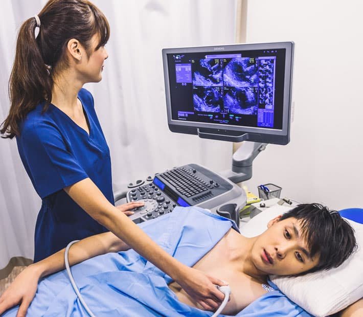 Echocardiography A healthcare professional in scrubs performs an echocardiogram on a young male patient lying on an examination table. The echocardiogram machine displays images of the heart on a monitor.