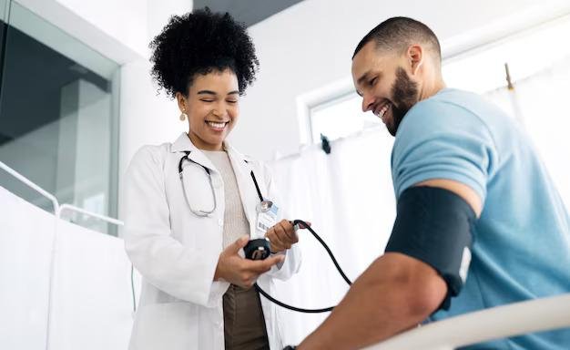 A full body checkup with Dr. Anil Batra​ A female doctor with curly hair, wearing a white coat and a stethoscope, is smiling as she checks the blood pressure of a male patient with short hair in a blue shirt. They are in a medical office with natural light coming in.